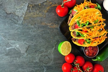 Hard shelled tacos with ground beef, lettuce, tomatoes and cheese. Top view, corner border on a dark background with copy space.
