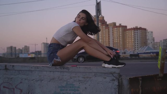cute sexy brunette smokes a cigarette. A woman sits on the side of the highway and smokes thoughtfully. Warm summer evening 4K