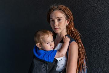 Young happy mom with baby son in ergo backpack travel together. Dark wall on the background. Concept of modern parents