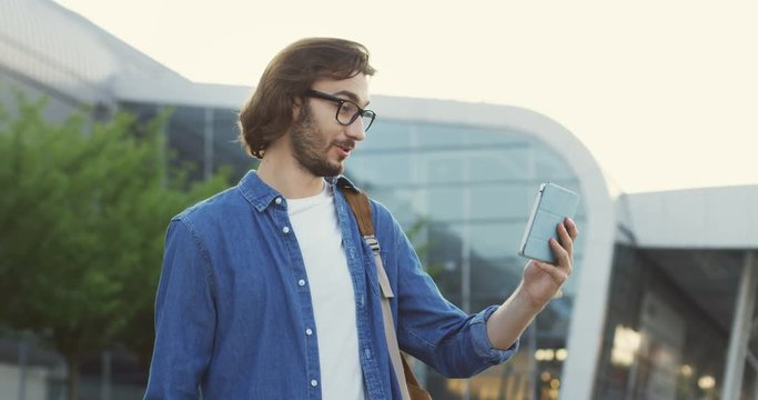 Young good looking man in glasses standing on the streen near big building like airport and having videochat on the tablet device.