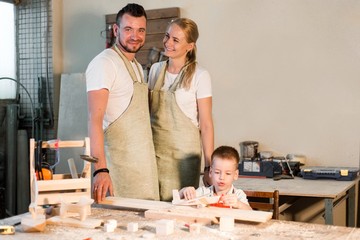 Happy family standing in carpentry workshop hugging each other. Son plays with a wooden statuette of an airplane