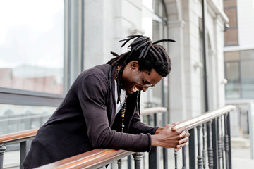 Happy African man with dreadlocks and glasses standing on the street leaning on the railing