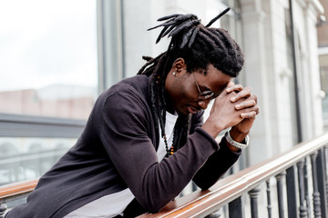 Thoughtful African man with dreadlocks and glasses standing on the street leaning on the railing