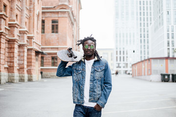 African man in denim jacket in sunglasses holding a skateboard standing on the street of the city - Powered by Adobe