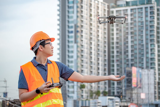 Young Asian engineer man flying drone over construction site. Using unmanned aerial vehicle (UAV) for land and building site survey in civil engineering project.
