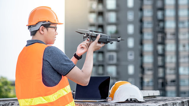 Asian engineer man working with drone and laptop computer at construction site. Using unmanned aerial vehicle (UAV) for land and building site survey in civil engineering project.