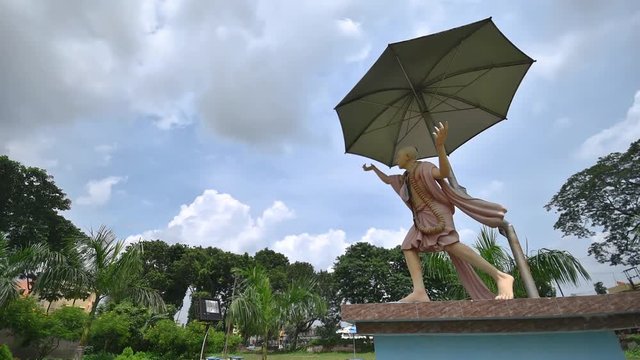 A timelapse of statue of Indian demi god Chaitanya Deb with umbrella and clouds in blue sky near a Hindu Temple