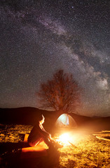 Camping night in mountains. Brightly lit by burning campfire dark silhouette of sitting young girl under dark starry sky with tourist tent and distant hills background. Tourism and travel concept.