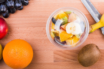 Fruit and berry salad in a plastic cup on a wooden board and ingredients - top view