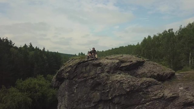 Drone view. Sniper woman with a sniper rifle sits on the rocks on the river. Cameta moving from object - sniper woman
