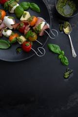 Apéro-Spiesse mit Tomaten, Käse, Schinken, Mozzarella und Pesto auf einem Teller auf einem alten dunklen Untergrund