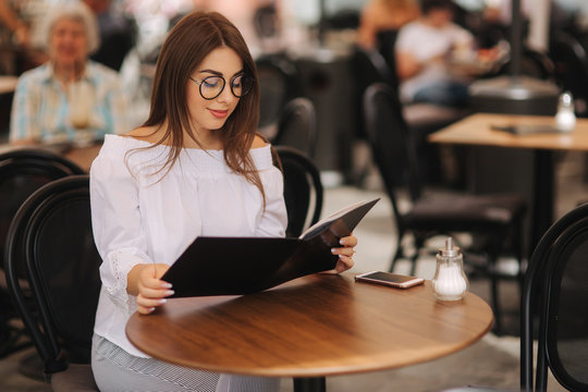 Stylish lady sits in cafe in centre of city and look at the menu. Sunny weather and codl drink. Girl in sunglasses chooses what to order