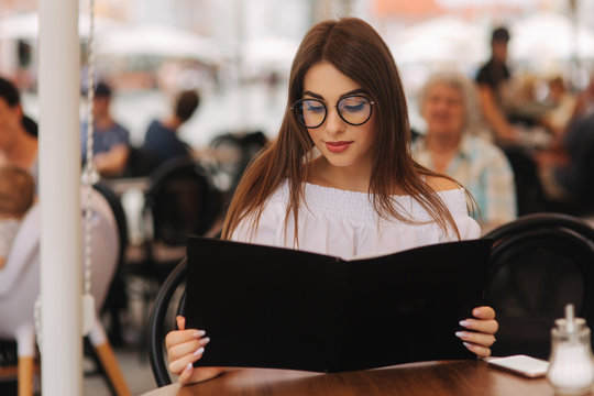 Stylish lady sits in cafe in centre of city and look at the menu. Sunny weather and codl drink. Girl in sunglasses chooses what to order