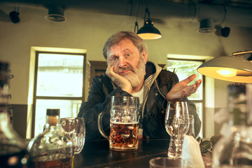 Sad senior bearded man drinking alcohol in pub and watching a sport program on TV. Enjoying my favorite teem and beer. Man with mug of beer sitting at table. Football or sport fan. Human emotions