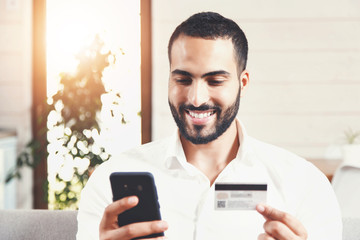Handsome bearded man in white shirt sitting on the sofa making online purchase via smartphone using credit card
