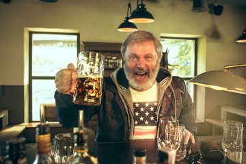 Senior bearded man drinking alcohol in pub and watching a sport program on TV. Enjoying my favorite teem and beer. Man with mug of beer sitting at table. Football or sport fan. Human emotions concept