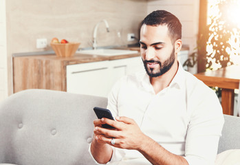 Sexy bearded man in white shirt sitting on the sofa and writting message on the smartphone, using app concept