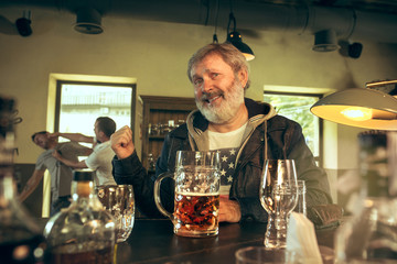 Senior bearded man drinking alcohol in pub and watching a sport program on TV. Enjoying beer. Man with mug of beer sitting at table. Football or sport fan. fight of fans in the background