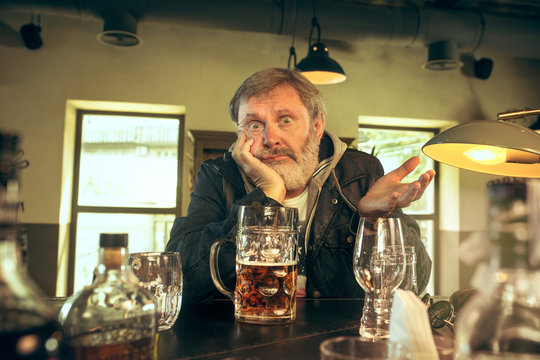 Sad senior bearded man drinking alcohol in pub and watching a sport program on TV. Enjoying my favorite teem and beer. Man with mug of beer sitting at table. Football or sport fan. Human emotions