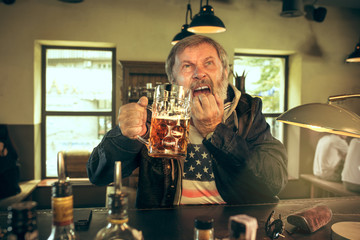 Sad senior bearded man drinking alcohol in pub and watching a sport program on TV. Enjoying my favorite teem and beer. Man with mug of beer sitting at table. Football or sport fan. Human emotions