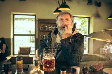 Sad senior bearded man drinking alcohol in pub and watching a sport program on TV. Enjoying my favorite teem and beer. Man with mug of beer sitting at table. Football or sport fan. Human emotions