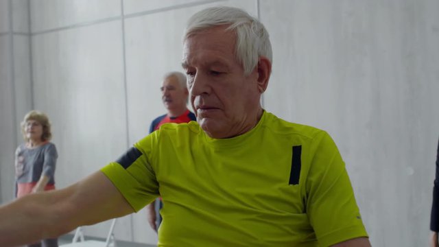 PAN with slowmo of group of senior people with grey hair doing cross-body arm stretch while warming up in fitness class