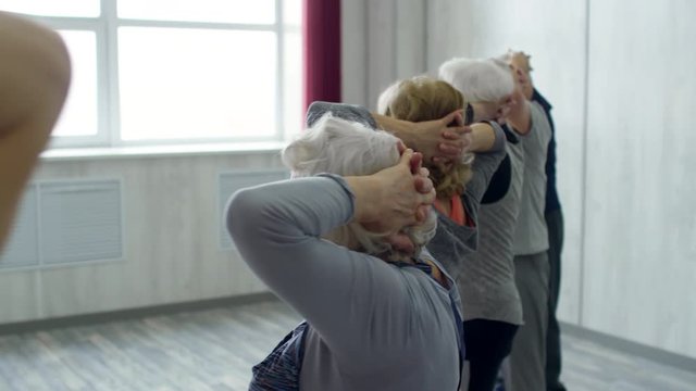 PAN of senior people with hands behind their heads standing in line and repeating after female fitness instructor doing torso twists during group class