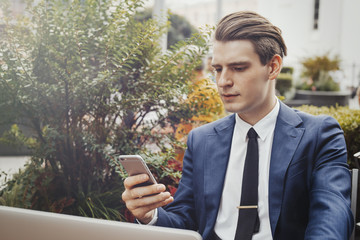 Young businessman holding mobile phone in hand and sitting next to green plant.