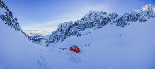 A red tent pitched up on snow in the middle of winter alpine landscape. Winter camping on snow. High alpine peaks, blue sky and bivi tent in winter alpine environment. Adventure and extreme alpinism. © Ondra