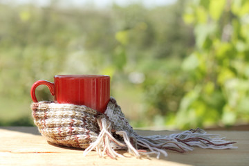 cozy rest on a sunny day/ red cup with a hot drink wrapped in scarf, stands on table in the garden terrace