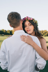 Obraz premium beautiful young bride in floral wreath hugging with groom and looking at camera in park