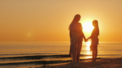 Fototapeta premium Mom and daughter are looking forward to a beautiful sunset over the sea. Silhouettes of a woman with a child near the water