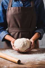 cropped shot of woman in apron holding raw dough for pizza in hands