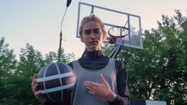 Attractive blonde female basketball player playing with ball and looking at camera, standing in park, daytime