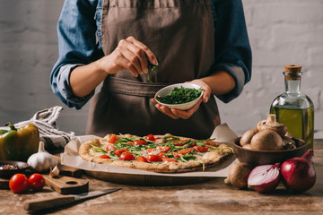 partial view of woman in apron pouring parsley on cooked pizza at wooden surface