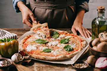 partial view of woman in apron taking piece of pizza on cutting board on wooden tabletop