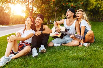 Company of friends having fun at sunset sitting on the grass, playing guitar, good summer mood, street party