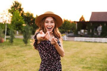 Photo of beautiful young woman 20s wearing dress and straw hat smiling, and inviting to follow her...
