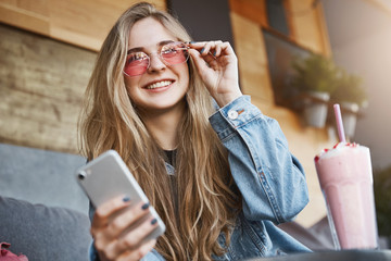 Attractive and joyful caucasian blond woman in trendy sunglasses, touching rim of eyewear and...