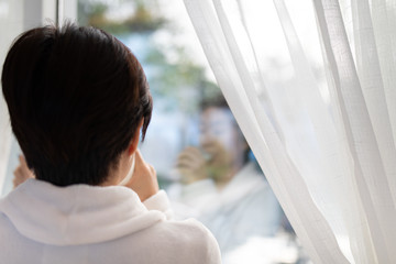 Rear view of Asian woman drinking a glass of milk in living room and looking outside window in the morning