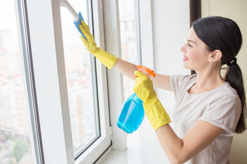 Nice and delightful girl stands in front of window and cleaning it with rag and blue liquid spray. Girl wears yellow gloves.