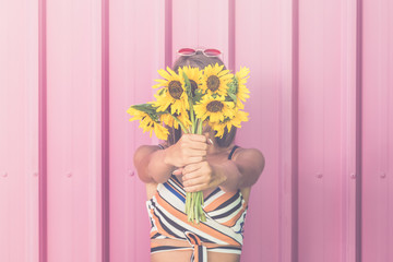 Fashionable woman covering her face with sunflowers against rose wall.