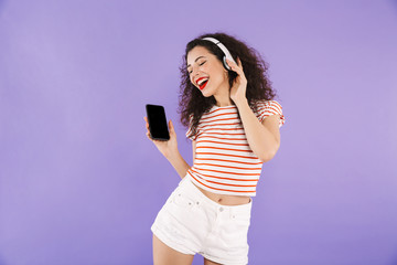 Naklejka premium Photo of adorable trendy woman with curly hair in summer wear listening to music via wireless headphones and holding cell phone, isolated over violet background in studio