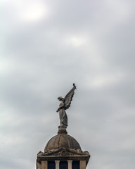 Fototapeta premium Angel sculpture and a magpie against the cloudy sky in Poblenou Cemetery. Cemetery of Poblenou is today home to incredible sculptures, haunting, yet beautiful.