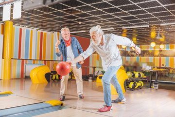 Interesting game. Joyful strong man throwing the bowling ball while playing together with his friend