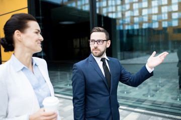 Bearded man in glasses and blue suit with showing hand gesture walking with woman in light clothes holding paper cup