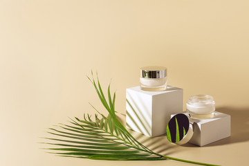 close up view of green palm leaf, facial and body creams in glass jars on white cubes on beige background