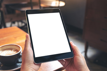 Mockup image of hands holding black tablet pc with blank white screen with coffee cup on wooden table in cafe