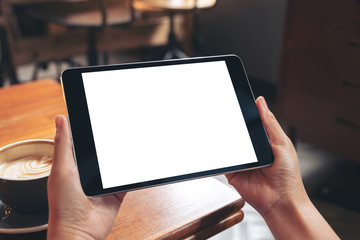 Mockup image of hands holding black tablet pc with blank white screen with coffee cup on wooden table in cafe