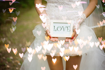 the bride in a short wedding dress and wearing gloves holds a plate with the word love in her hands with tot of hearts bokeh